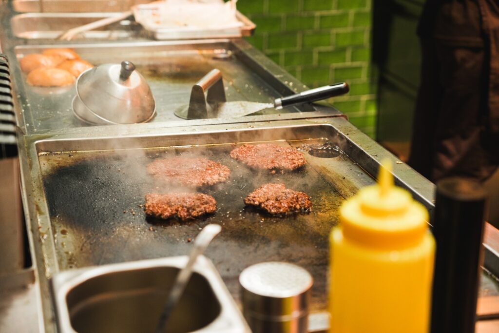 Drei Burger-Patties beim Grillen im Port Island Hamburg mit Dampf, Spachtel und klassischer Kücheneinrichtung. Port Island Hamburg